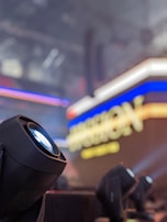 A black stage light is prominently featured in the foreground, with a blurred background displaying an illuminated sign with vibrant blue and yellow text. The scene is indoors, likely in an event or concert setting.