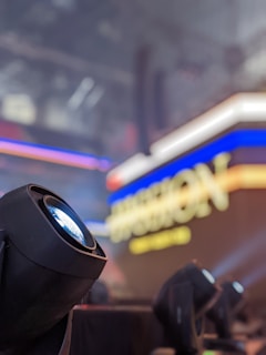 A black stage light is prominently featured in the foreground, with a blurred background displaying an illuminated sign with vibrant blue and yellow text. The scene is indoors, likely in an event or concert setting.