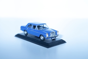 A miniature blue vintage car model is placed on a black display stand against a plain, light blue background. The model appears detailed, capturing the essence of a classic car design with prominent headlights and chrome accents.