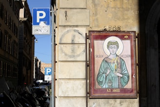 A painting of a religious figure dressed in robes holding a syringe and a bowl. The figure has a halo and is surrounded by Latin text. The image is mounted on an exterior wall of a building. Nearby, a parking sign is visible with a blue P and an additional sign indicating restrictions for bicycles.