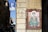 A painting of a religious figure dressed in robes holding a syringe and a bowl. The figure has a halo and is surrounded by Latin text. The image is mounted on an exterior wall of a building. Nearby, a parking sign is visible with a blue P and an additional sign indicating restrictions for bicycles.