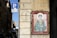 A painting of a religious figure dressed in robes holding a syringe and a bowl. The figure has a halo and is surrounded by Latin text. The image is mounted on an exterior wall of a building. Nearby, a parking sign is visible with a blue P and an additional sign indicating restrictions for bicycles.