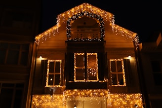 A house adorned with numerous warm glowing string lights, creating a festive and cozy atmosphere. The lights outline the structure's roof and windows, with additional lights forming an arch above a central balcony. The scene is captured at night, enhancing the brightness and warmth of the lights against the dark sky.