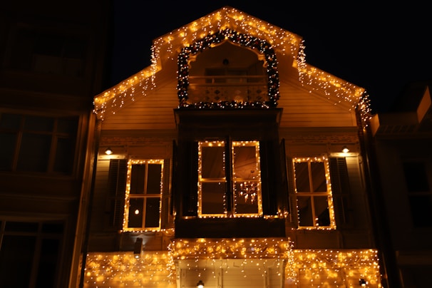 A cozy residential home adorned with warm white string lights outlining the windows and roof, glowing softly against the night sky.