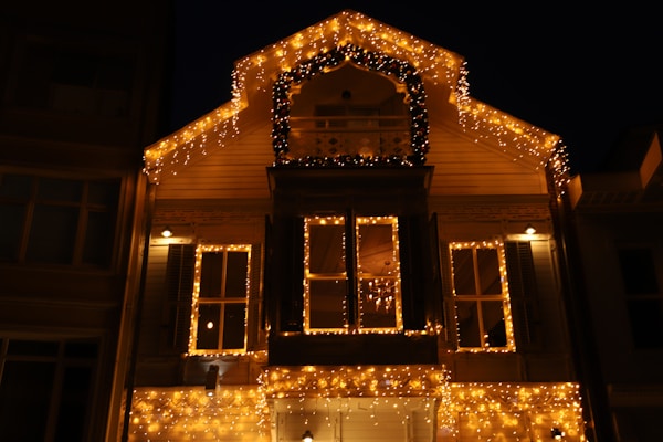 A house adorned with numerous warm glowing string lights, creating a festive and cozy atmosphere. The lights outline the structure's roof and windows, with additional lights forming an arch above a central balcony. The scene is captured at night, enhancing the brightness and warmth of the lights against the dark sky.