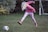 A child with blonde hair wearing a pink hoodie, colorful leggings, and pink boots is actively kicking a black-and-white soccer ball on a grassy area. In the background, there is a brick wall and a small soccer goal post.