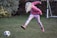 A child with blonde hair wearing a pink hoodie, colorful leggings, and pink boots is actively kicking a black-and-white soccer ball on a grassy area. In the background, there is a brick wall and a small soccer goal post.