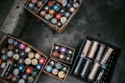 Several cardboard boxes and a crate contain an assortment of spray paint cans in various colors, organized neatly on a concrete floor.