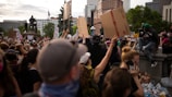 A large crowd of people is gathered at a protest, holding up various signs. A statue is visible in the background, surrounded by tall buildings and trees. One person is sitting on a ledge, appearing to capture the scene with a camera.