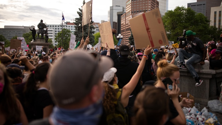 A large crowd of people is gathered at a protest, holding up various signs. A statue is visible in the background, surrounded by tall buildings and trees. One person is sitting on a ledge, appearing to capture the scene with a camera.
