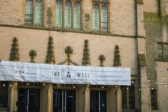 An ornate building facade made of stone with intricate carvings between tall, narrow windows featuring stained glass. A banner with the words 'The Well' is displayed prominently, along with social media handles and a website address. There are decorative string lights hanging across the entrance.
