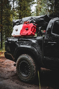 A rugged, off-road vehicle with the logo 'TRD 4x4 Sport' is parked in a forest setting. The vehicle is equipped with large, all-terrain tires and carries camping and off-road gear on its roof rack, including red and white fuel canisters and a black cargo bag labeled '23ZERO'. The trees and dirt road suggest a remote, adventurous location.