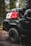A rugged, off-road vehicle with the logo 'TRD 4x4 Sport' is parked in a forest setting. The vehicle is equipped with large, all-terrain tires and carries camping and off-road gear on its roof rack, including red and white fuel canisters and a black cargo bag labeled '23ZERO'. The trees and dirt road suggest a remote, adventurous location.