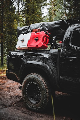 A rugged, off-road vehicle with the logo 'TRD 4x4 Sport' is parked in a forest setting. The vehicle is equipped with large, all-terrain tires and carries camping and off-road gear on its roof rack, including red and white fuel canisters and a black cargo bag labeled '23ZERO'. The trees and dirt road suggest a remote, adventurous location.