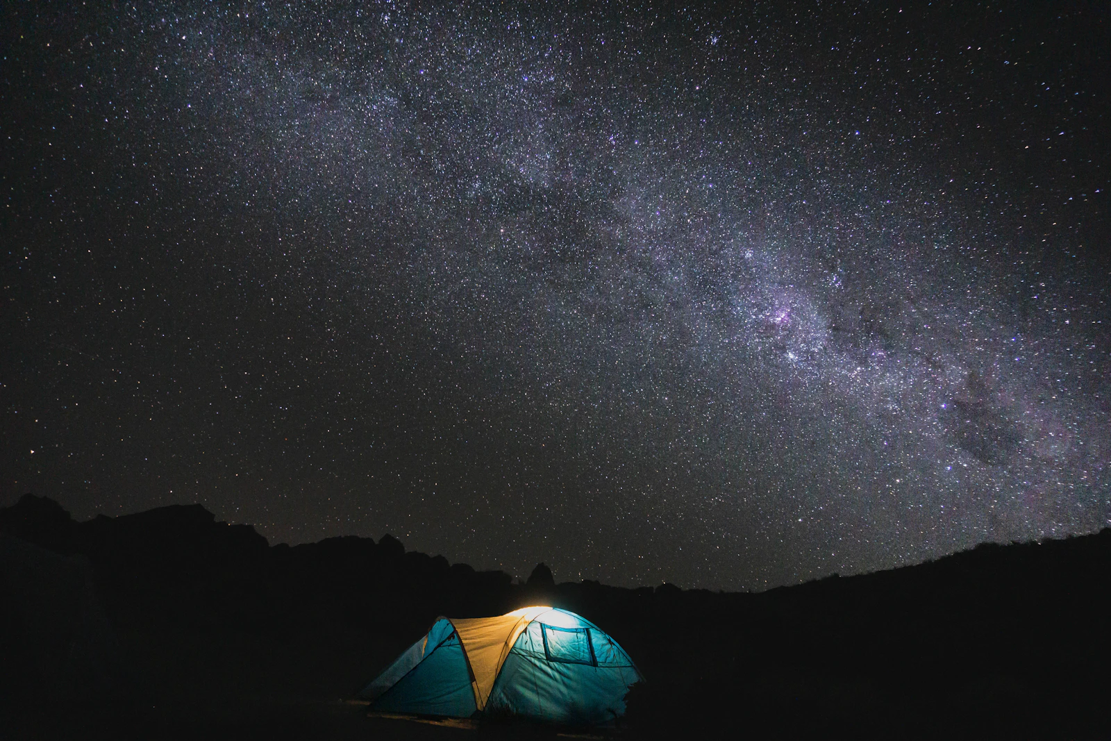 A tent glowing beneath a dense field of stars and the Milky Way over the dark prairie