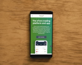 A smartphone displaying an advertisement for an online trading platform called eToro. The screen shows a green background with text promoting the platform as a leading social trading platform. A button labeled 'Get Started' is visible, along with a partial view of a mobile app screen below.