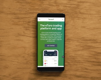 A smartphone displaying an advertisement for an online trading platform called eToro. The screen shows a green background with text promoting the platform as a leading social trading platform. A button labeled 'Get Started' is visible, along with a partial view of a mobile app screen below.