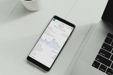 A smartphone is placed on a white desk showing a stock trading app with a line graph and financial details. Next to the phone is the keyboard of a laptop, and a white cup is in the top left corner.