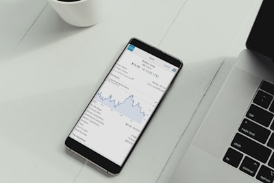 A smartphone is placed on a white desk showing a stock trading app with a line graph and financial details. Next to the phone is the keyboard of a laptop, and a white cup is in the top left corner.