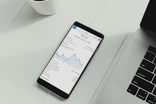 A smartphone is placed on a white desk showing a stock trading app with a line graph and financial details. Next to the phone is the keyboard of a laptop, and a white cup is in the top left corner.