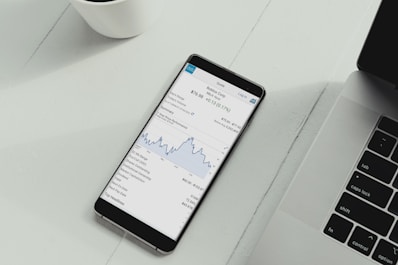 A smartphone is placed on a white desk showing a stock trading app with a line graph and financial details. Next to the phone is the keyboard of a laptop, and a white cup is in the top left corner.