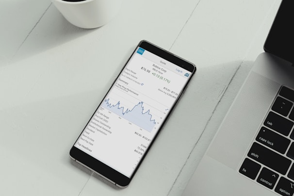 A smartphone is placed on a white desk showing a stock trading app with a line graph and financial details. Next to the phone is the keyboard of a laptop, and a white cup is in the top left corner.
