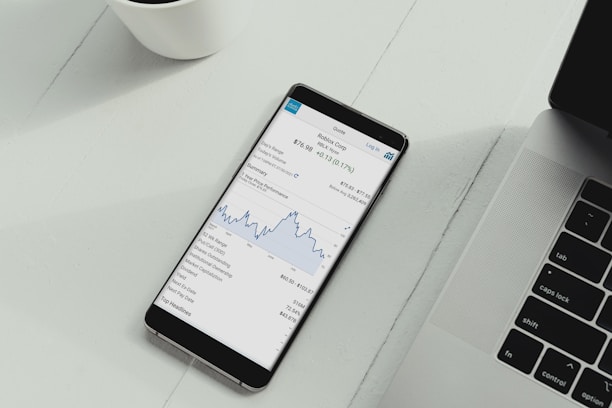 A smartphone is placed on a white desk showing a stock trading app with a line graph and financial details. Next to the phone is the keyboard of a laptop, and a white cup is in the top left corner.