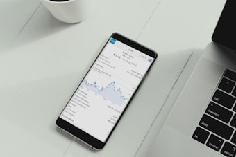 A smartphone is placed on a white desk showing a stock trading app with a line graph and financial details. Next to the phone is the keyboard of a laptop, and a white cup is in the top left corner.