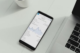 A smartphone is placed on a white desk showing a stock trading app with a line graph and financial details. Next to the phone is the keyboard of a laptop, and a white cup is in the top left corner.