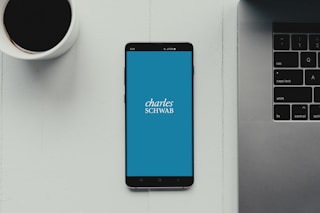 A modern workspace setup featuring a smartphone with the Charles Schwab logo displayed on the screen. It rests on a light-colored table beside a cup of black coffee and a part of a laptop keyboard.