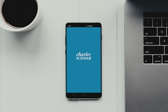 A modern workspace setup featuring a smartphone with the Charles Schwab logo displayed on the screen. It rests on a light-colored table beside a cup of black coffee and a part of a laptop keyboard.