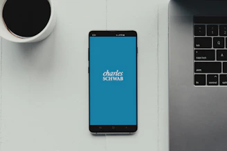 A modern workspace setup featuring a smartphone with the Charles Schwab logo displayed on the screen. It rests on a light-colored table beside a cup of black coffee and a part of a laptop keyboard.