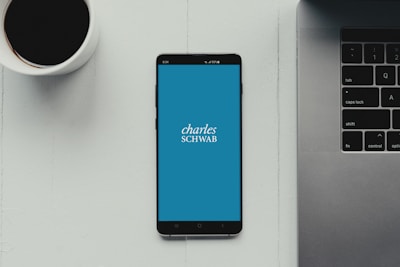A modern workspace setup featuring a smartphone with the Charles Schwab logo displayed on the screen. It rests on a light-colored table beside a cup of black coffee and a part of a laptop keyboard.