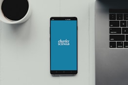 A modern workspace setup featuring a smartphone with the Charles Schwab logo displayed on the screen. It rests on a light-colored table beside a cup of black coffee and a part of a laptop keyboard.