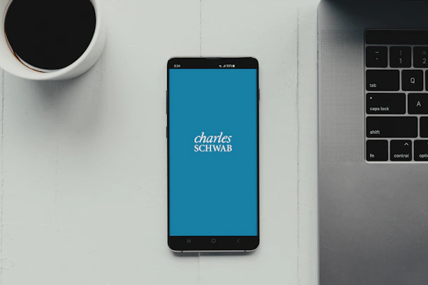 A modern workspace setup featuring a smartphone with the Charles Schwab logo displayed on the screen. It rests on a light-colored table beside a cup of black coffee and a part of a laptop keyboard.