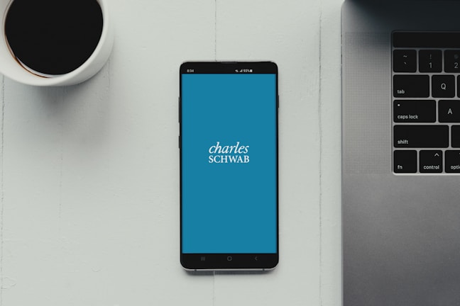 A modern workspace setup featuring a smartphone with the Charles Schwab logo displayed on the screen. It rests on a light-colored table beside a cup of black coffee and a part of a laptop keyboard.