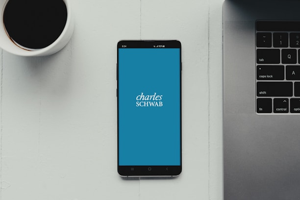 A modern workspace setup featuring a smartphone with the Charles Schwab logo displayed on the screen. It rests on a light-colored table beside a cup of black coffee and a part of a laptop keyboard.