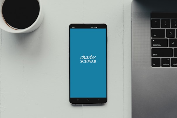 A modern workspace setup featuring a smartphone with the Charles Schwab logo displayed on the screen. It rests on a light-colored table beside a cup of black coffee and a part of a laptop keyboard.