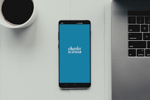 A modern workspace setup featuring a smartphone with the Charles Schwab logo displayed on the screen. It rests on a light-colored table beside a cup of black coffee and a part of a laptop keyboard.