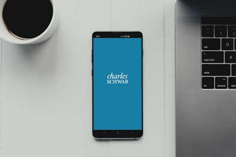 A modern workspace setup featuring a smartphone with the Charles Schwab logo displayed on the screen. It rests on a light-colored table beside a cup of black coffee and a part of a laptop keyboard.