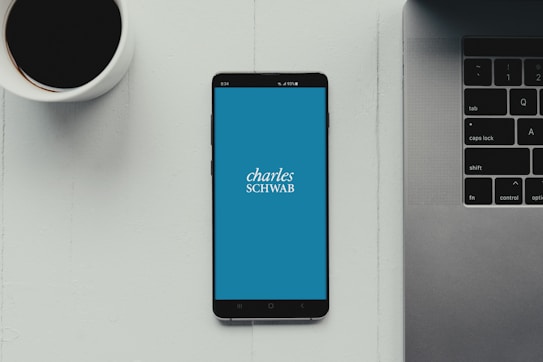 A modern workspace setup featuring a smartphone with the Charles Schwab logo displayed on the screen. It rests on a light-colored table beside a cup of black coffee and a part of a laptop keyboard.