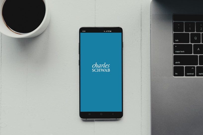 A modern workspace setup featuring a smartphone with the Charles Schwab logo displayed on the screen. It rests on a light-colored table beside a cup of black coffee and a part of a laptop keyboard.