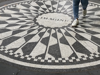 An intricate mosaic pattern on the ground with the word 'IMAGINE' at the center. A person wearing blue jeans and white sneakers is stepping across the design.
