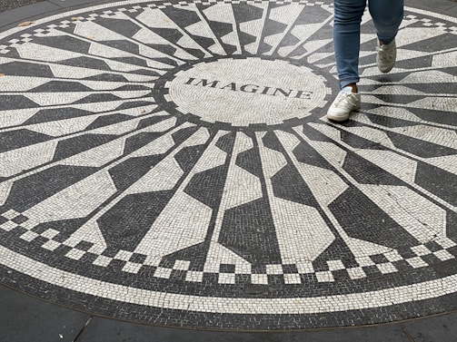 An intricate mosaic pattern on the ground with the word 'IMAGINE' at the center. A person wearing blue jeans and white sneakers is stepping across the design.