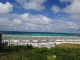 A scenic beach view with white sand and thatched umbrellas lining the shore. The turquoise ocean stretches to the horizon, under a sky scattered with fluffy clouds. Lush greenery borders the beach area, adding a touch of vibrant color to the scene.