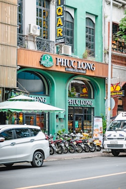 A city street scene with a row of parked motorbikes in front of a coffee and tea shop called Phuc Long. The facade of the shop is painted in green with large glass windows. Above the shop entrance is a brown sign with white letters displaying the shop's name. Part of a white SUV and a van with decorative decals are visible on the street. There's a green awning above the entrance, and the building's upper floors have large windows and signs for a lounge.