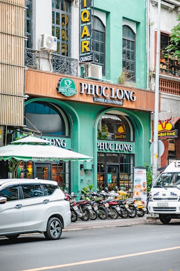 A city street scene with a row of parked motorbikes in front of a coffee and tea shop called Phuc Long. The facade of the shop is painted in green with large glass windows. Above the shop entrance is a brown sign with white letters displaying the shop's name. Part of a white SUV and a van with decorative decals are visible on the street. There's a green awning above the entrance, and the building's upper floors have large windows and signs for a lounge.