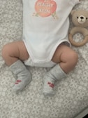 A baby wearing a white onesie with a peach design and the words 'PEACHY KEEN' written on it. The baby has gray socks with red text that reads 'MY FIRST XMAS'. A knitted teddy bear rattle is next to the baby, resting on a patterned surface.