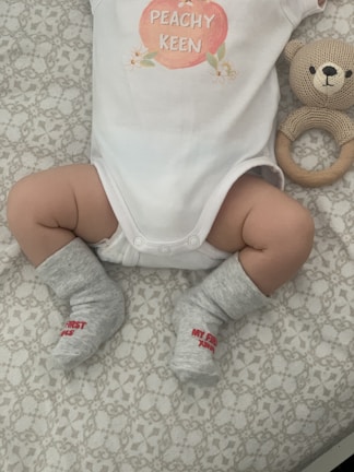 A baby wearing a white onesie with a peach design and the words 'PEACHY KEEN' written on it. The baby has gray socks with red text that reads 'MY FIRST XMAS'. A knitted teddy bear rattle is next to the baby, resting on a patterned surface.