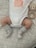 A baby wearing a white onesie with a peach design and the words 'PEACHY KEEN' written on it. The baby has gray socks with red text that reads 'MY FIRST XMAS'. A knitted teddy bear rattle is next to the baby, resting on a patterned surface.